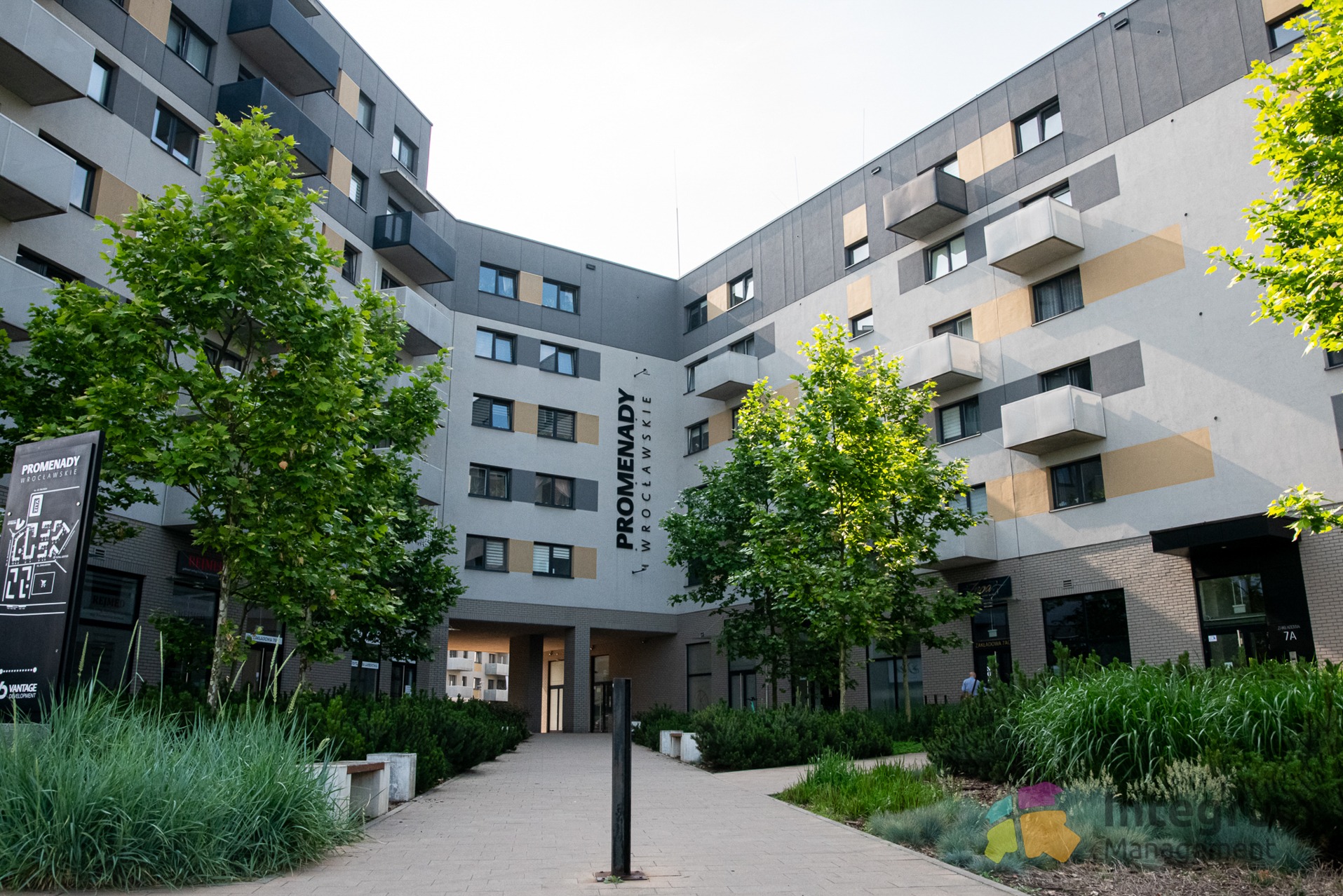 Nowoczesny budynek mieszkalny 'Promenady Wrocławskie' z zielenią i chodnikiem, widziany z perspektywy przechodnia. Architektura z balkonami i elementami dekoracyjnymi.