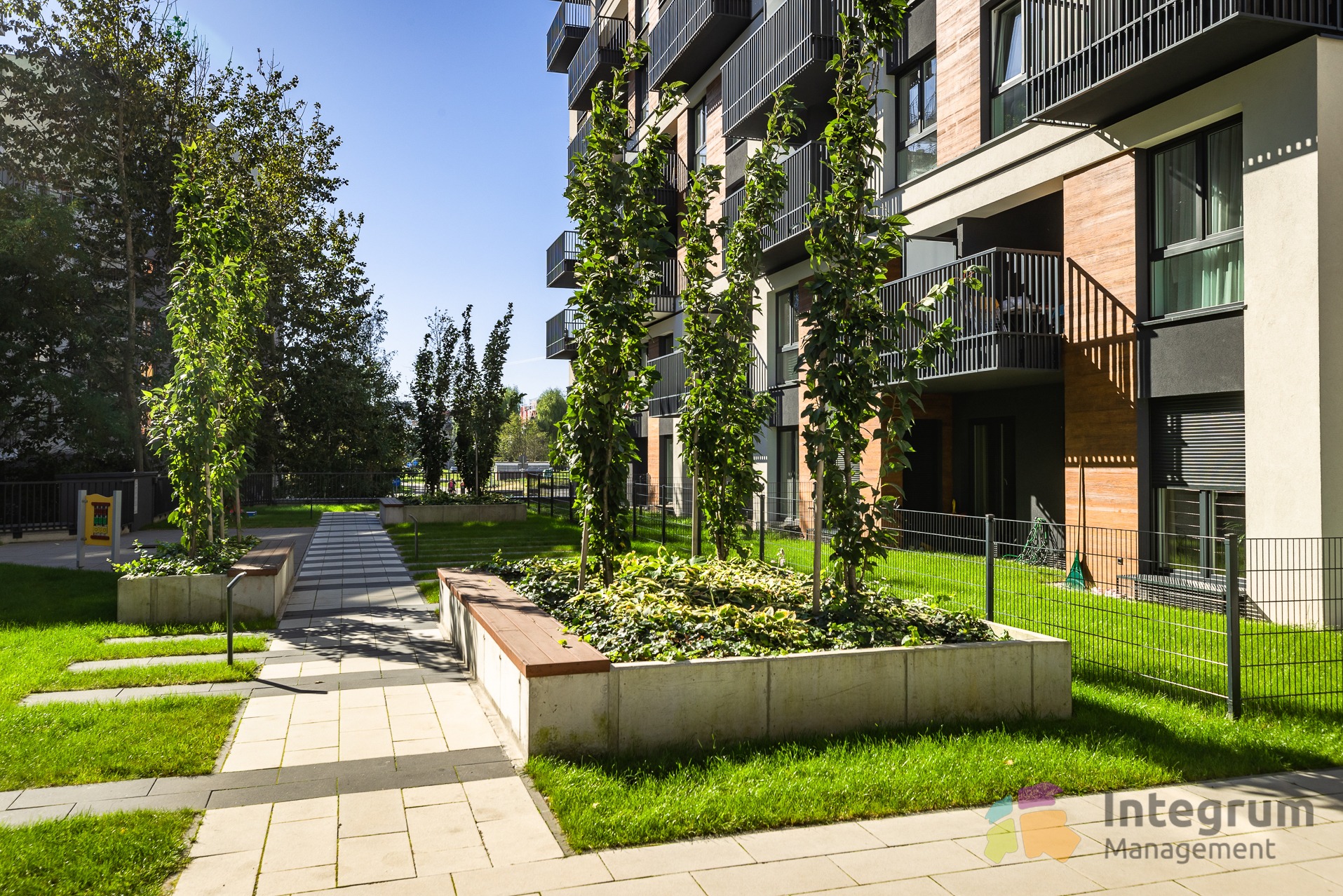 Nowoczesny budynek wielorodzinny z balkonami i zielenią: drzewa w donicach, trawnik, ścieżka z kostki. Elementy małej architektury.