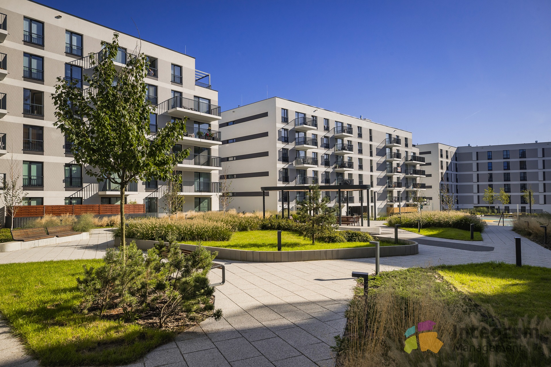 Nowoczesne osiedle mieszkaniowe z zadbanym dziedzińcem, zielenią i alejkami. Budynki w odcieniach szarości z balkonami. Słoneczny dzień, błękitne niebo.