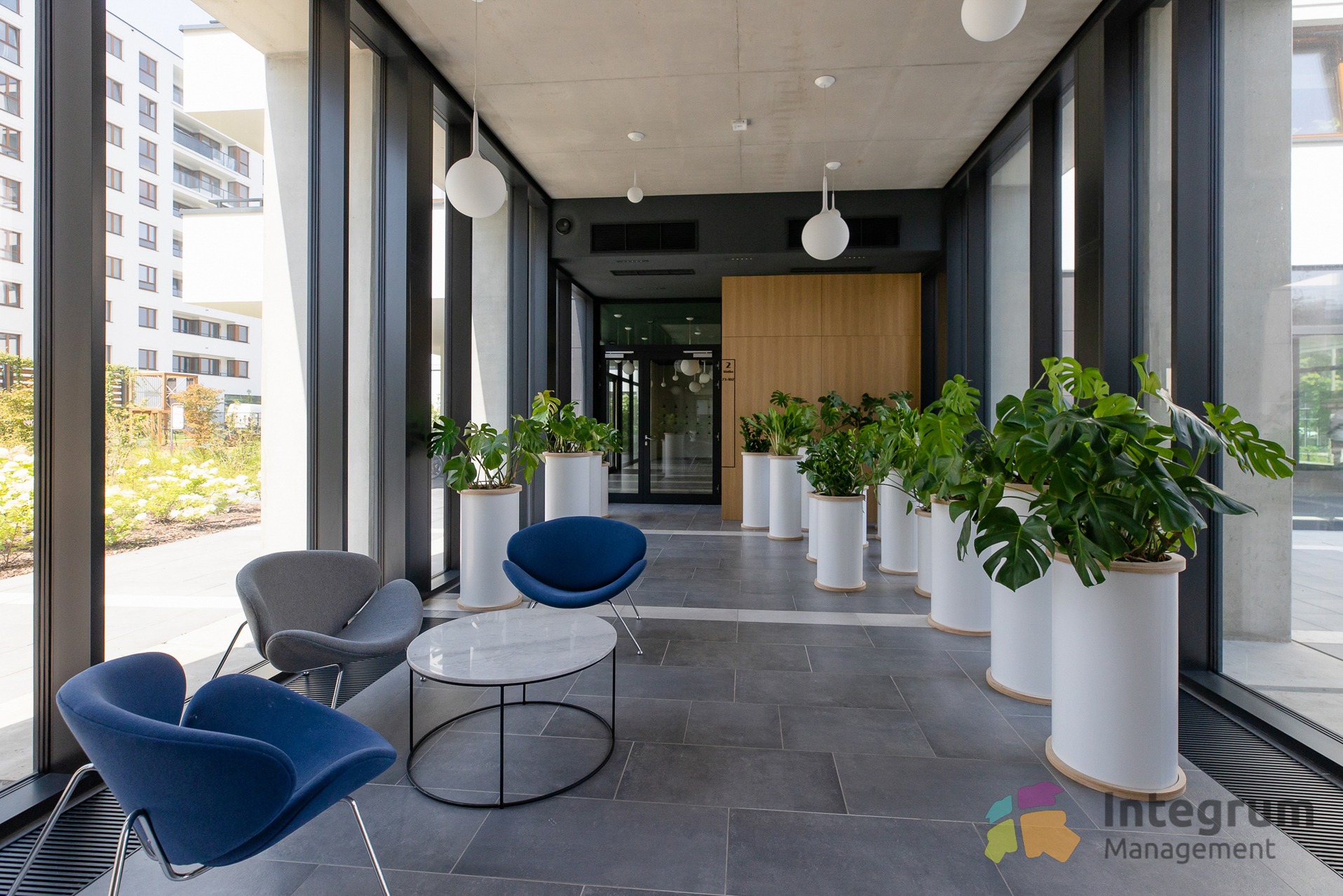 Nowoczesne wnętrze holu apartamentowca z roślinami w białych donicach, niebieskimi krzesłami i marmurowym stolikiem. Widok na zewnątrz przez duże okna.