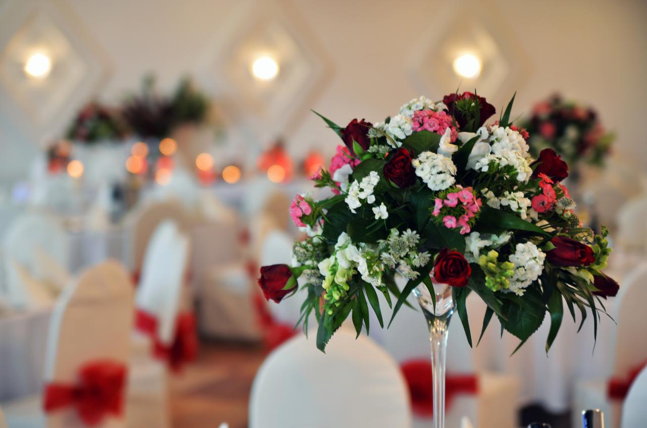 Dekoracja kwiatowa ze świeżych róż, białych i różowych kwiatów w wysokim, smukłym wazonie na stole. W tle sala bankietowa z białymi krzesłami i dekoracjami.