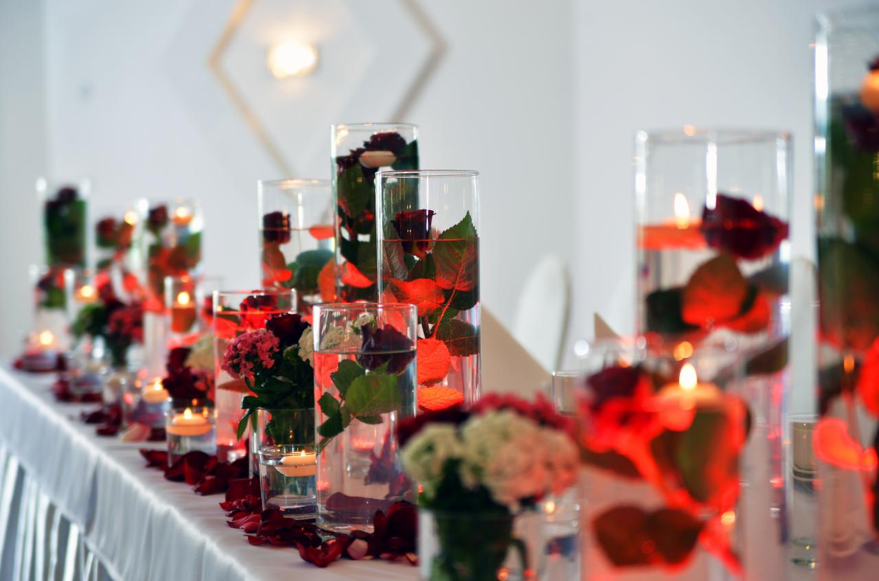Dekoracja stołu weselnego z kwiatami i świecami w szklanych naczyniach, płatki róż rozsypane na białym obrusie. Romantyczny nastrój, ciepłe oświetlenie.