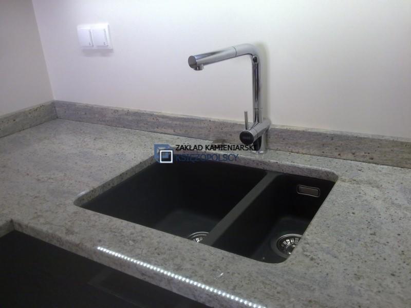 Szary blat granitowy z dwukomorowym, czarnym zlewem i chromowaną baterią. Pod blatem widoczna listwa LED. W tle biała ściana z włącznikiem światła.