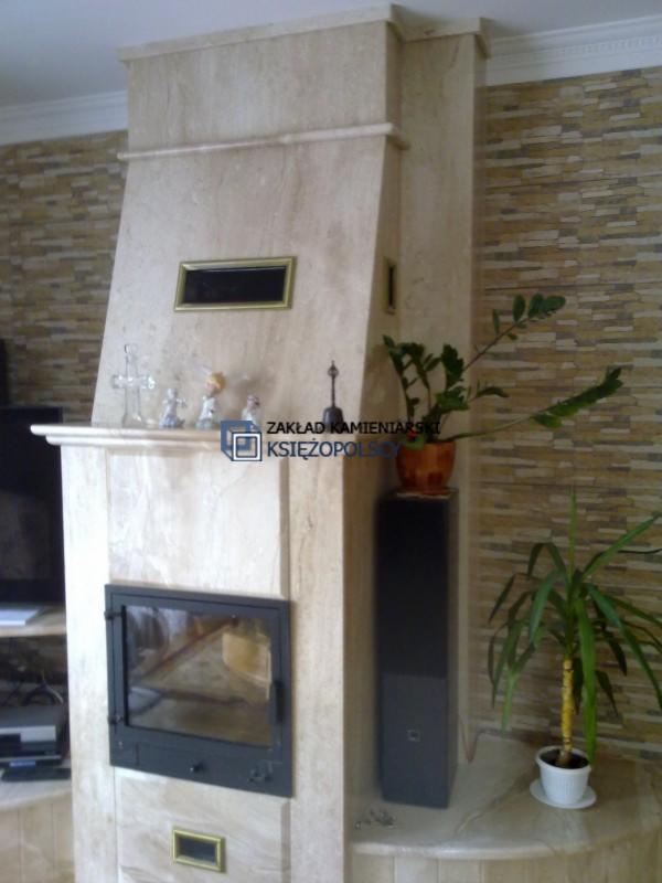 Obudowa kominkowa z jasnego kamienia z czarnym wkładem, ozdobnymi figurkami i roślinami doniczkowymi po bokach, na tle ściany z dekoracyjnej cegły.
