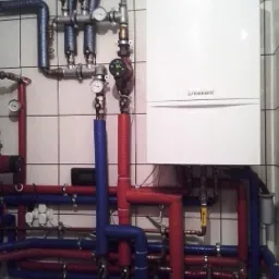 Biały piec Vaillant na tle kafelkowanej ściany, z widoczną instalacją hydrauliczną z rur w izolacji czerwonej i niebieskiej oraz manometrami.