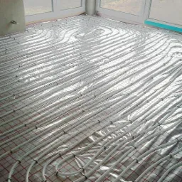 Białe rury ogrzewania podłogowego ułożone na folii z czerwoną siatką, przed wylaniem posadzki. Widok na jasne, duże okno w tle.