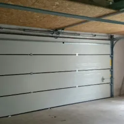 Zamknięta, segmentowa brama garażowa w jasnym kolorze, z widocznym mechanizmem otwierania i szynami montażowymi w surowym garażu z betonową posadzką.