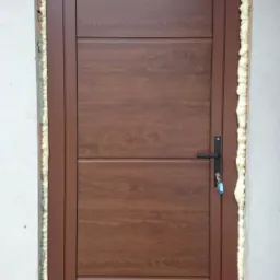 Brązowe drzwi zewnętrzne z czterema panelami o drewnopodobnej fakturze, zamontowane z użyciem pianki montażowej, widoczne klucze w zamku.