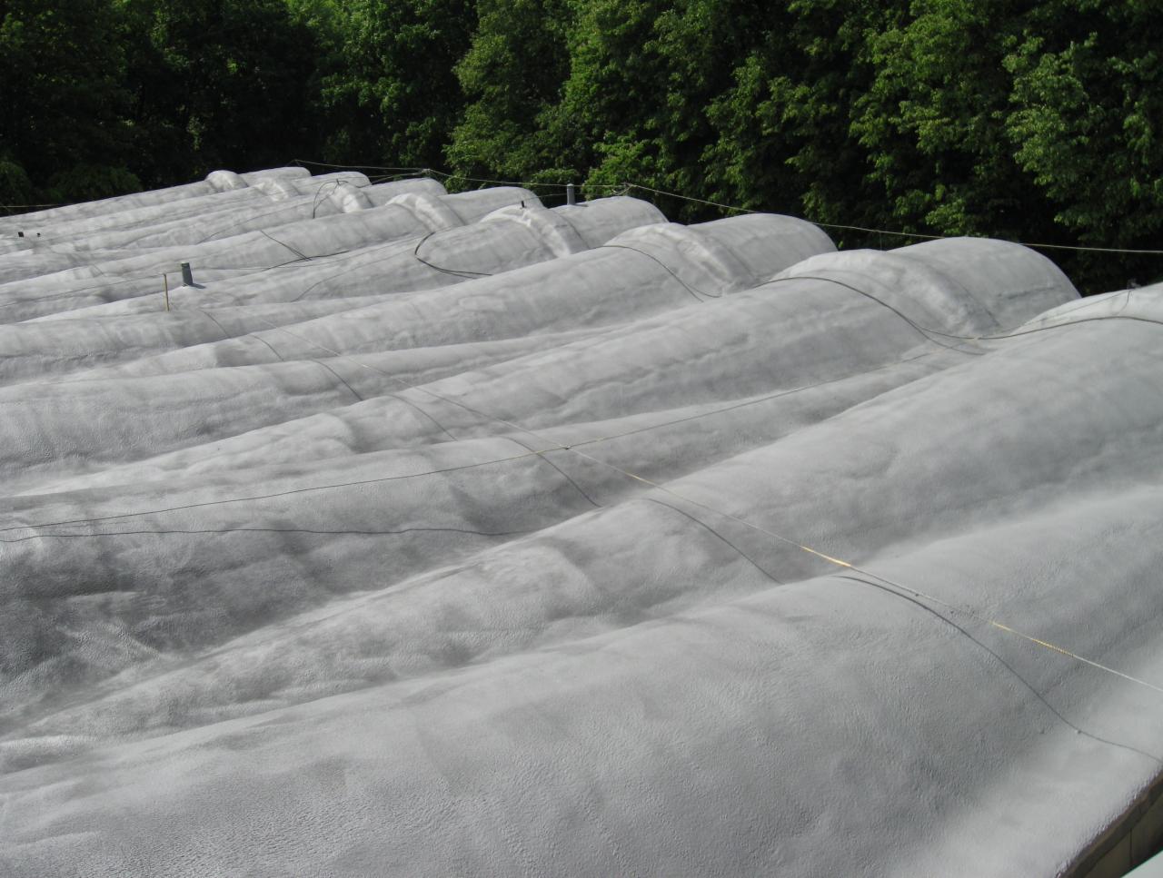 Szary, pofalowany dach po aplikacji izolacji, widok z góry, zieleń drzew w tle. Tekstura pianki widoczna z bliska.
