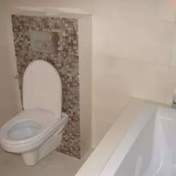 Biała, podwieszana toaleta z otwartą deską, zamontowana na ścianie wyłożonej mozaiką w odcieniach brązu i beżu, obok fragmentu białej wanny i chromowanej baterii.