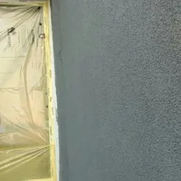 Szara ściana z fakturą baranka, zabezpieczone okno folią malarską, widoczny fragment podłogi zabezpieczony kartonem.