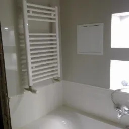 Biały grzejnik drabinkowy zamontowany nad wanną w łazience z panelami ściennymi, widoczna klapka rewizyjna i fragment baterii wannowej.