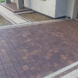 Ułożona kostka brukowa w Chrzanowie, z obrzeżami z jasnego kamienia. Widoczny podjazd i chodnik prowadzący do domu, wykonane z kostki w odcieniach brązu.