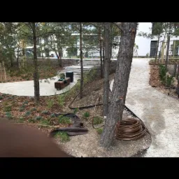 Ścieżka z utwardzoną nawierzchnią w parku, obok drzewa zwinięty wąż do nawadniania, w tle ławka i schody.