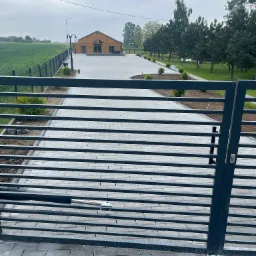 Widok na posesję przez metalową bramę: szara kostka brukowa na podjeździe, drewniany budynek w tle, zielony trawnik i pole po lewej.