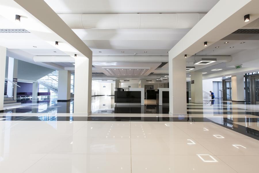 Przestronne, jasne wnętrze holu z błyszczącą podłogą w szachownicę, białymi kolumnami i podwieszanym sufitem z oświetleniem. Osoba sprzątająca w tle.