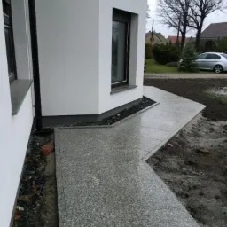 Szara nawierzchnia z drobnego kamienia przy białym budynku z czarnymi oknami. Widoczne drzewa w tle. Krawężnik z czarnych kamyk&oacute;w.