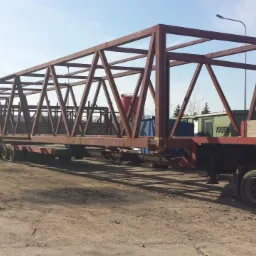 Metalowa konstrukcja mostu przewożona na platformie transportowej z wieloma osiami, na placu składowym.