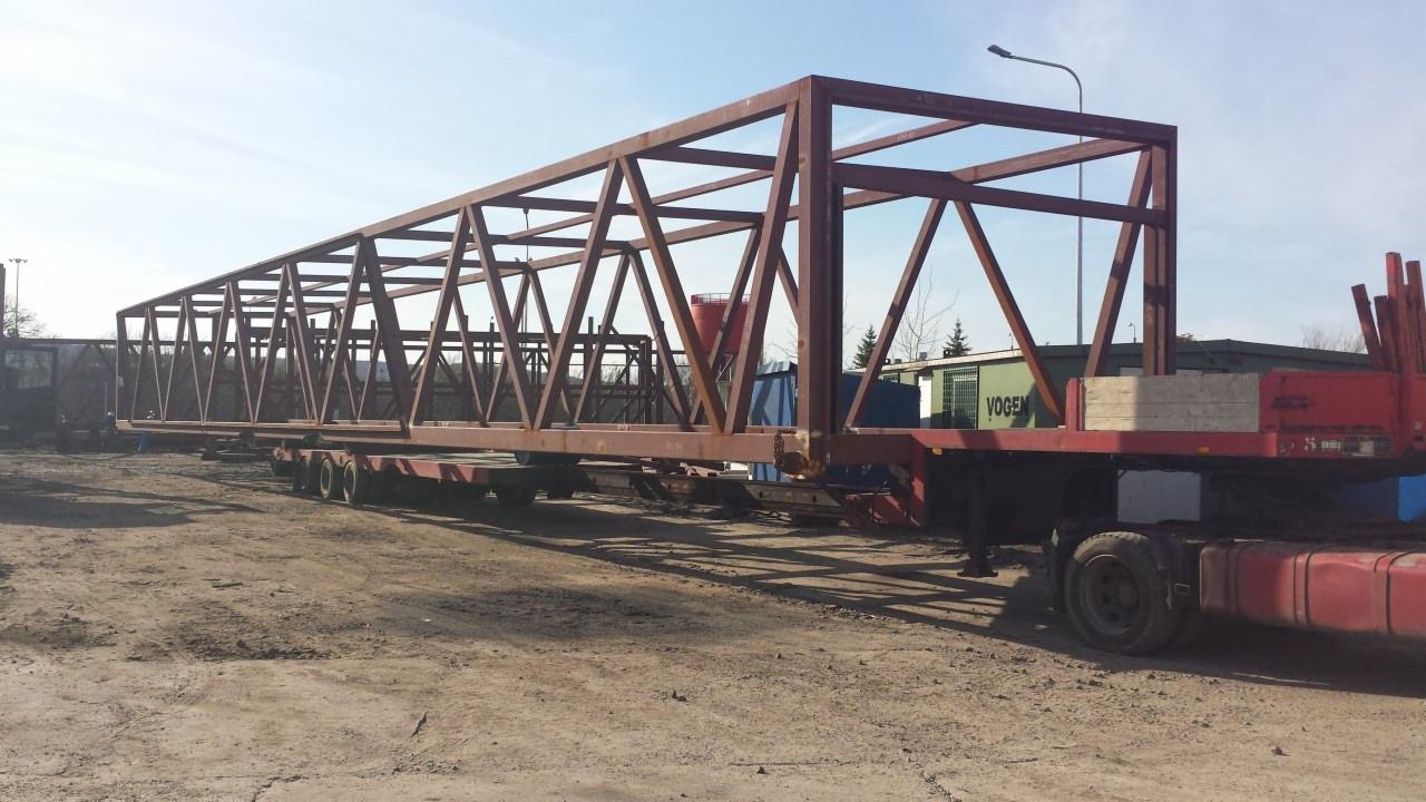 Metalowa konstrukcja mostu przewożona na platformie transportowej z wieloma osiami, na placu składowym.