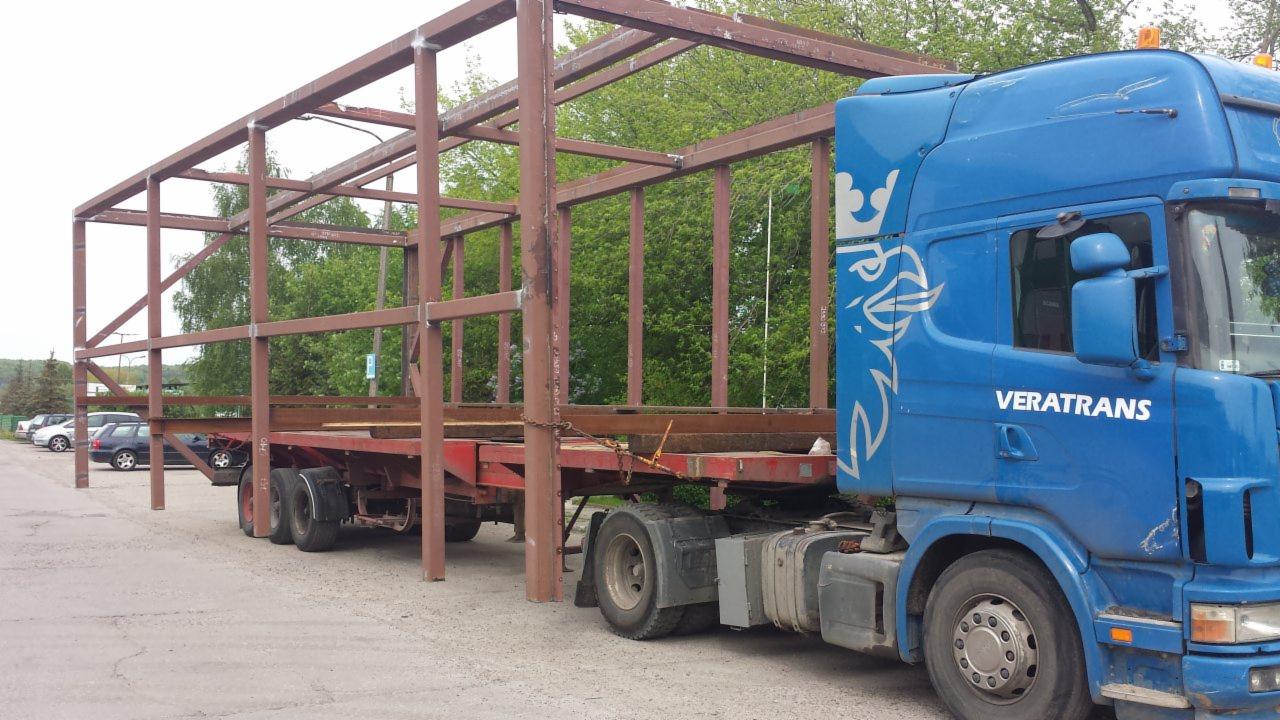 Niebieska ciężarówka firmy Veratrans z naczepą przewożąca brązową, metalową konstrukcję szkieletową na tle zieleni.