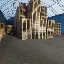 Magazyn z wysokimi stosami drewnianych palet euro pod zadaszeniem z metalową konstrukcją i niebieskim tłem