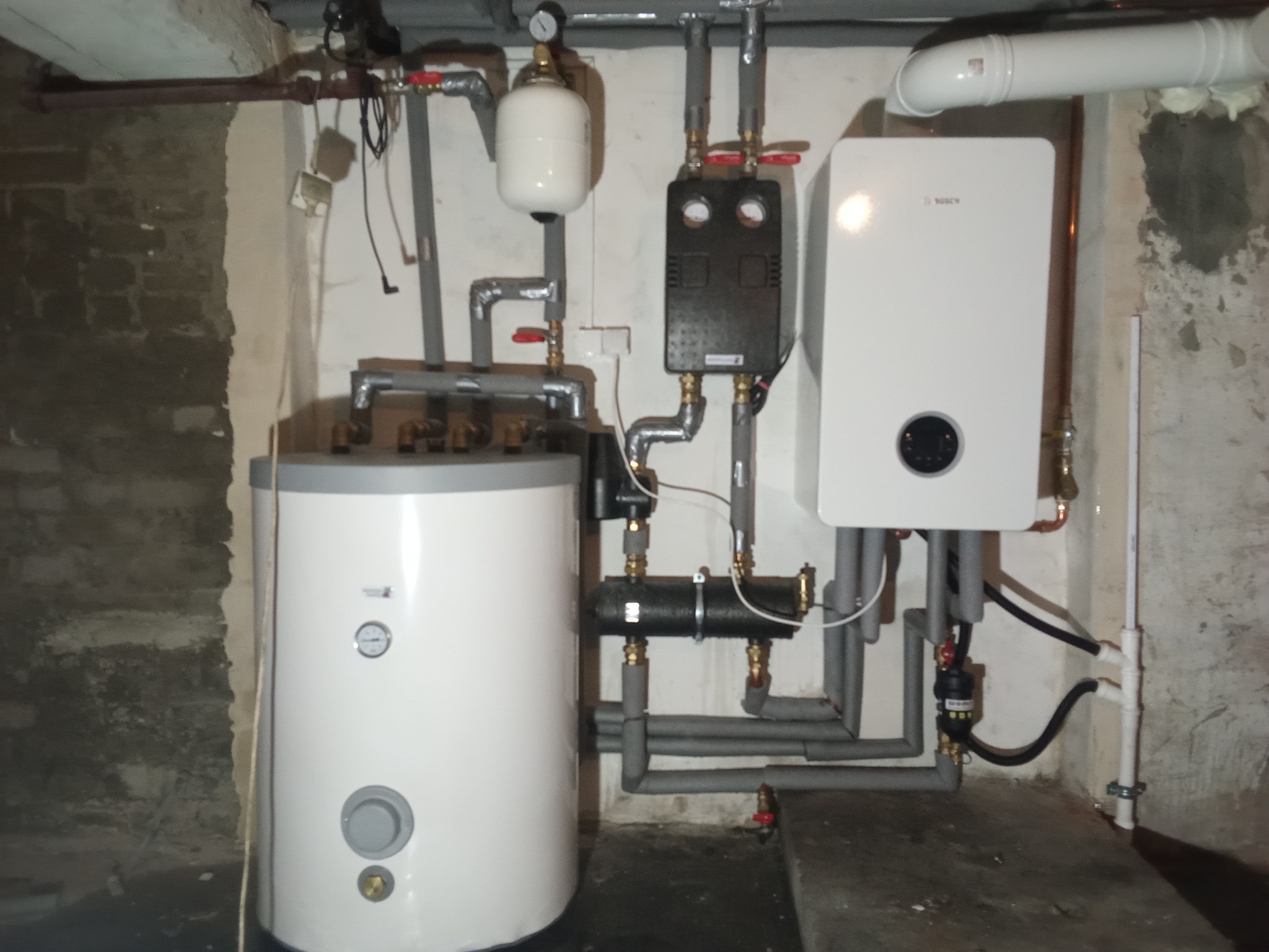 Kotłownia w piwnicy: biały bojler z manometrem, wiszący kocioł grzewczy marki Bosch, czarna skrzynka sterująca, rury instalacji grzewczej z zaworami, szara izolacja termiczna na rurach, surowe...