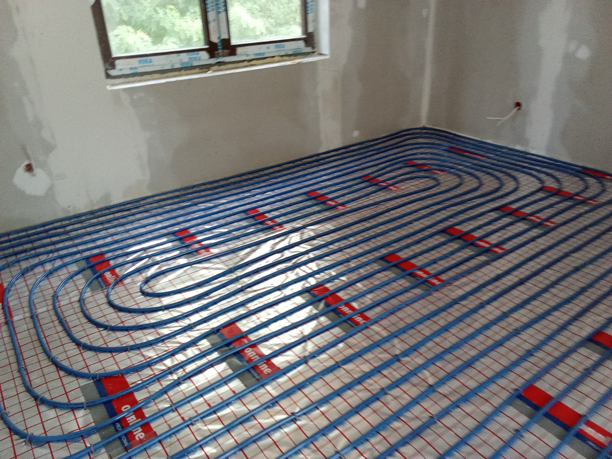 Niebieskie rury ogrzewania podłogowego ułożone w pętle na srebrnej folii z czerwoną kratką, widoczne szare ściany i okno w surowym stanie.