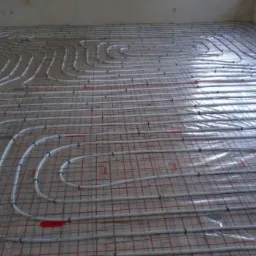 Widok z góry na ułożone rury ogrzewania podłogowego na siatce zbrojeniowej. Białe rury tworzą spiralny wzór na podłodze, gotowe do zalania betonem.