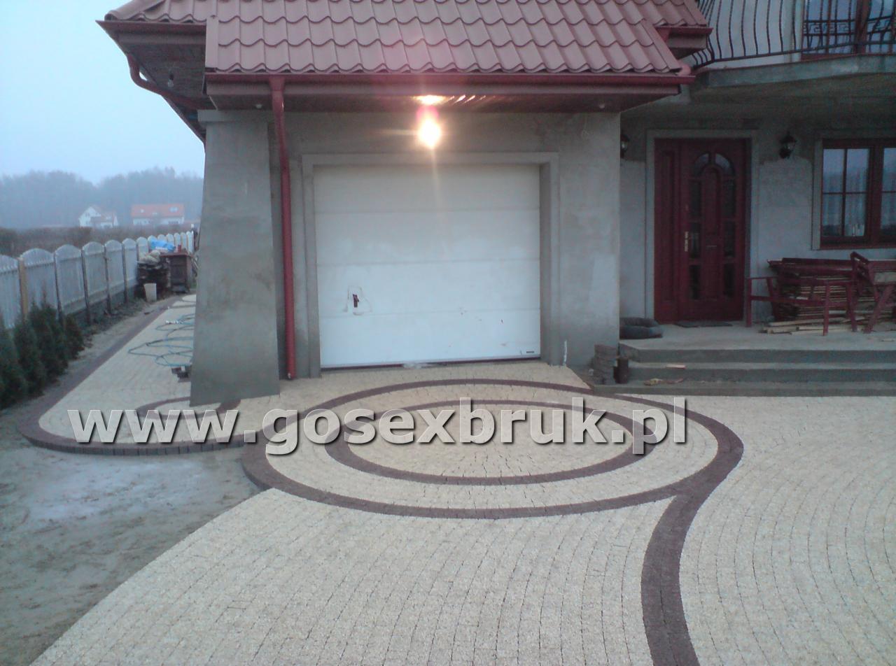 Ułożona kostka brukowa w okrągły wzór przed garażem i wejściem do domu. Jasna kostka z ciemnobrązowymi obramowaniami. Widoczna brama garażowa i schody wejściowe.