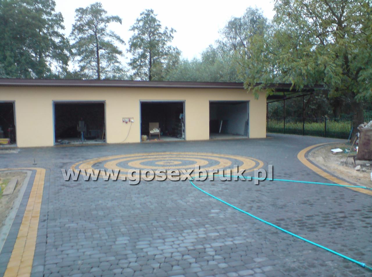 Ułożona kostka brukowa w kształcie koncentrycznych okręgów przed budynkiem garażowym. Widoczny wąż ogrodowy. Na kostce znak wodny z adresem strony internetowej.