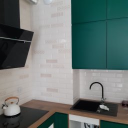 Monter- bud - Fragment nowej kuchni z zielonymi szafkami, białymi płytkami z delikatnym wzorem, czarnym zlewem i płytą grzewczą oraz białym czajnikiem.