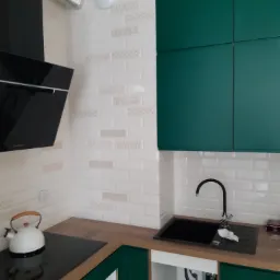 Fragment nowej kuchni z zielonymi szafkami, białymi płytkami z delikatnym wzorem, czarnym zlewem i płytą grzewczą oraz białym czajnikiem.