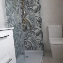 Nowoczesna łazienka po remoncie: kabina prysznicowa z deszczownicą i wzorzystymi płytkami w motywie roślinnym, biała umywalka z szafką i toaleta typu kompakt.