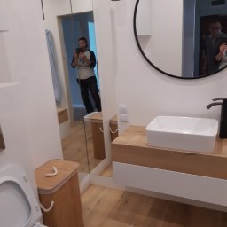 Monter- bud - Nowoczesna łazienka z drewnianymi akcentami, białą umywalką nablatową i okrągłym lustrem; widoczne odbicie osoby robiącej zdjęcie w lustrze.