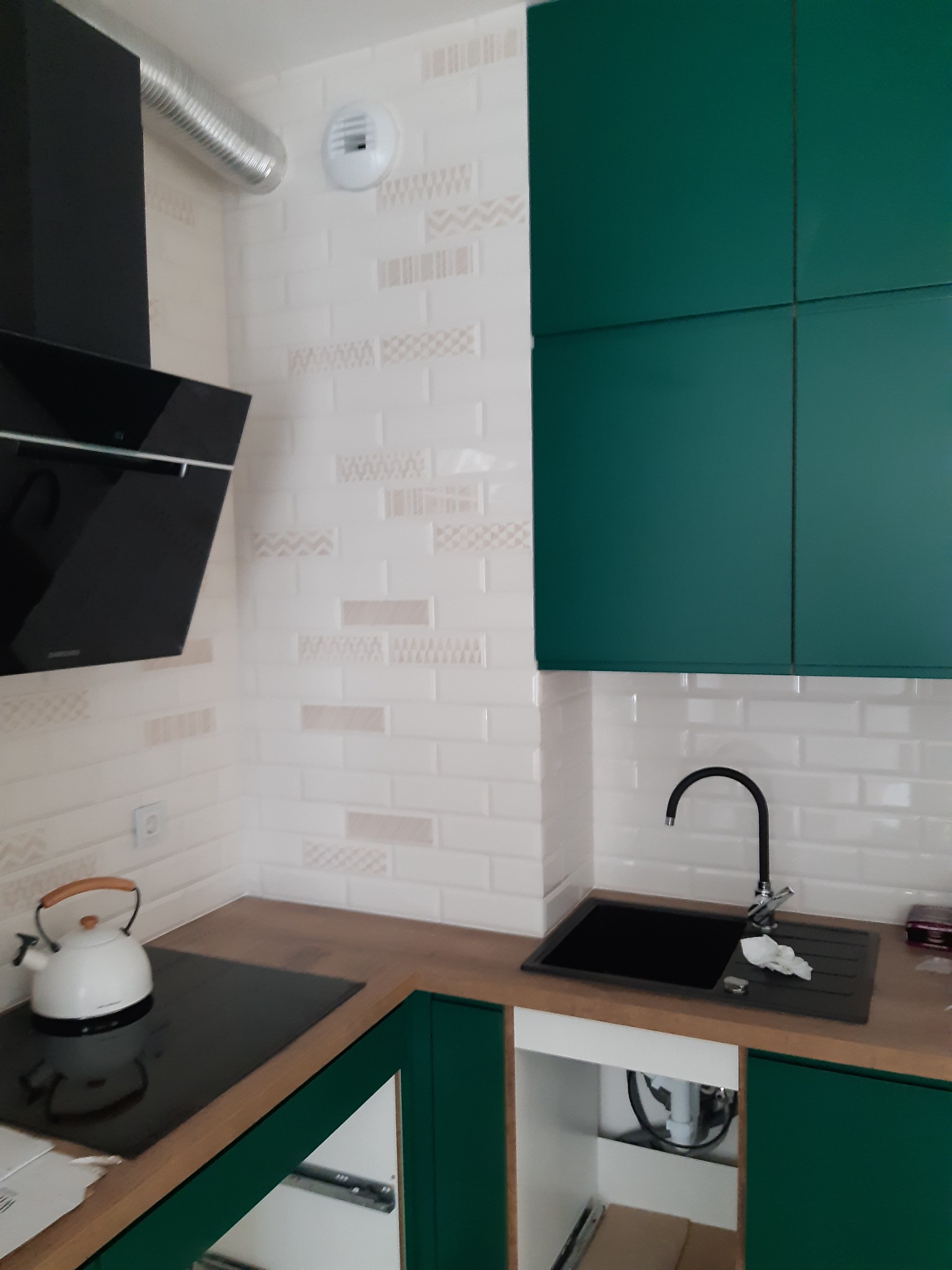 Fragment nowej kuchni z zielonymi szafkami, białymi płytkami z delikatnym wzorem, czarnym zlewem i płytą grzewczą oraz białym czajnikiem.