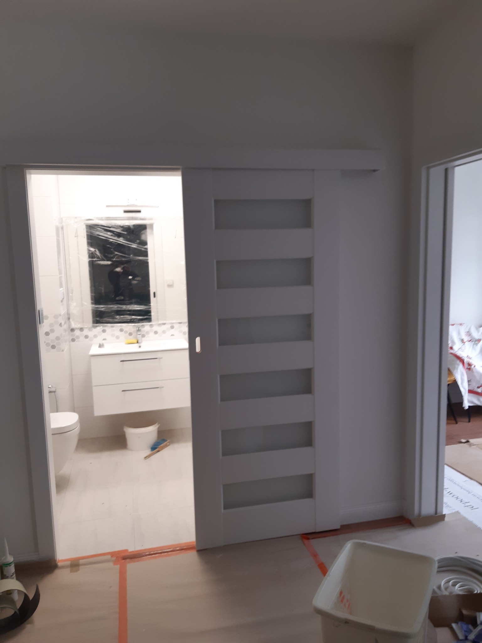 Białe przesuwne drzwi z szybami w trakcie montażu, widok na wyremontowaną łazienkę z umywalką, toaletą i lustrem zabezpieczonym folią, widoczne ślady prac wykończeniowych.