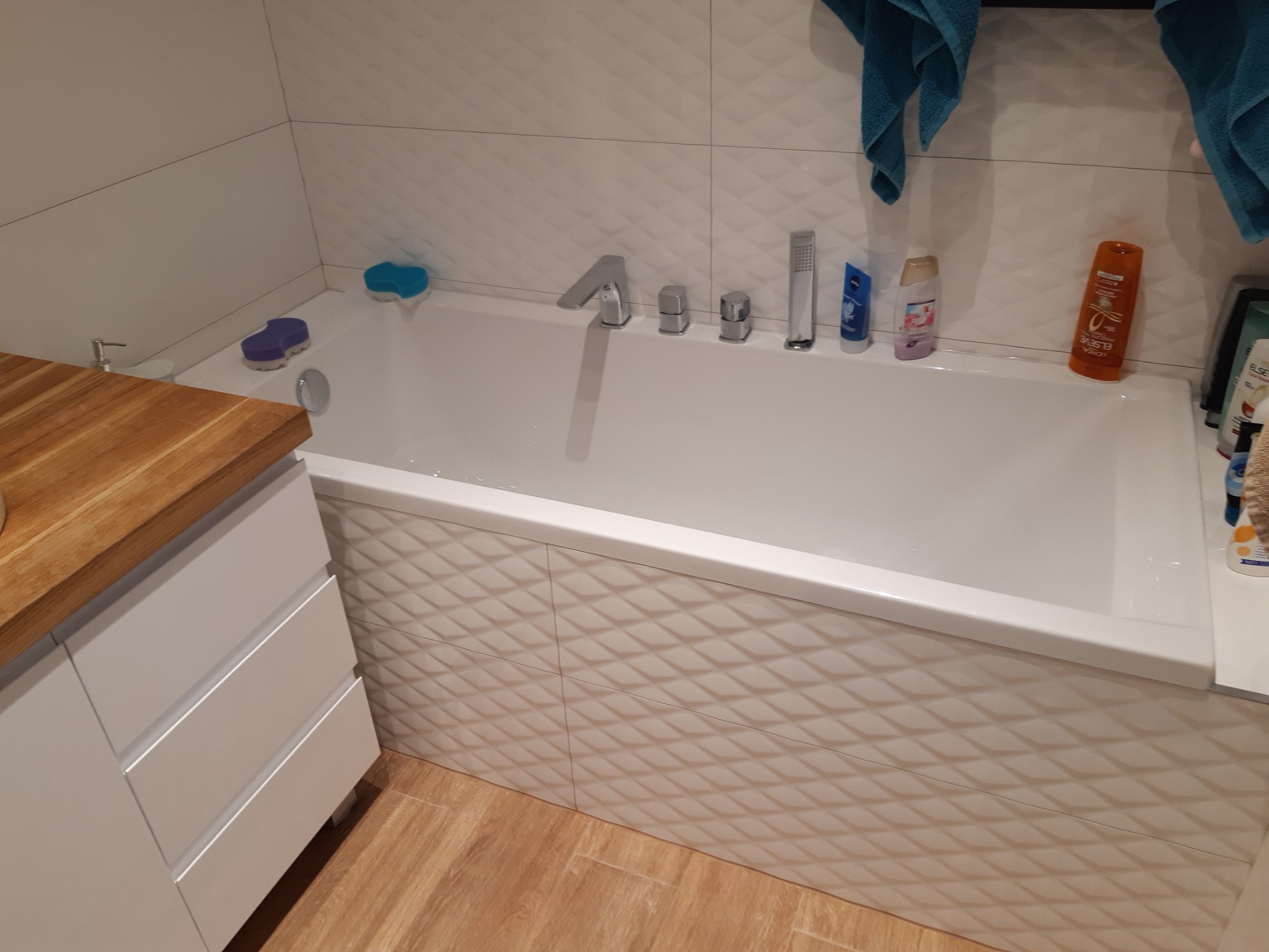 Nowoczesna łazienka z białą wanną obudowaną płytkami 3D, drewniany blat przy umywalce i błękitne ręczniki.