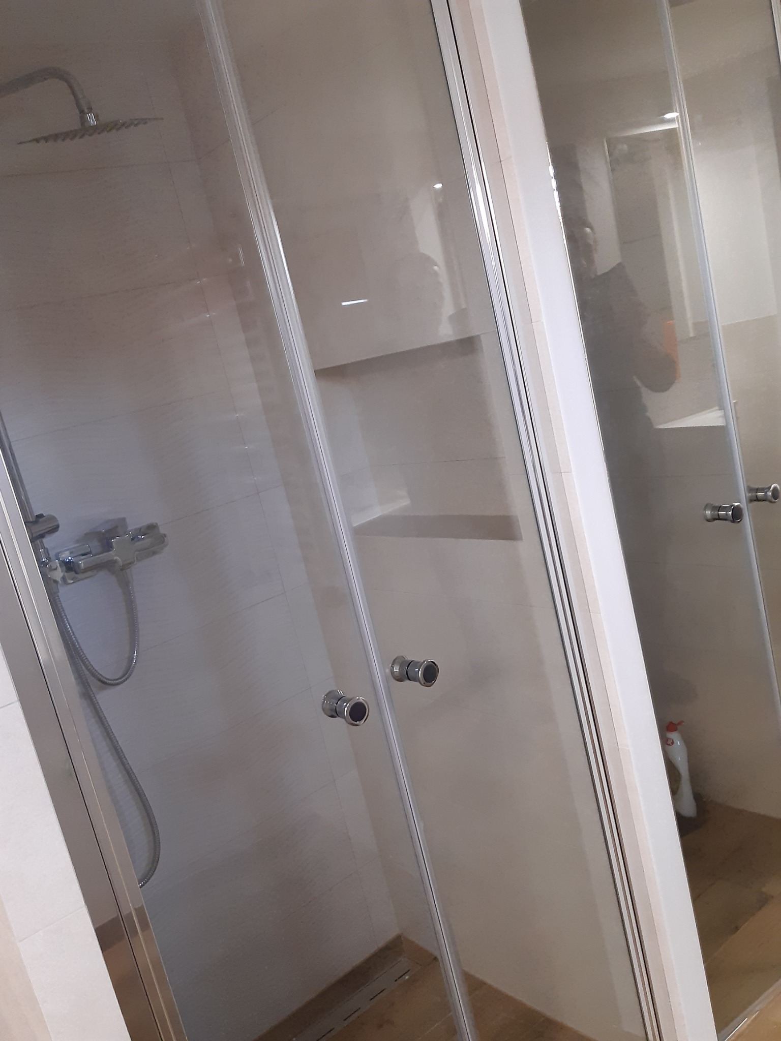 Kabina prysznicowa z chromowaną armaturą i szklanymi drzwiami, widoczne półki w ścianie i butelka z płynem czyszczącym w rogu.