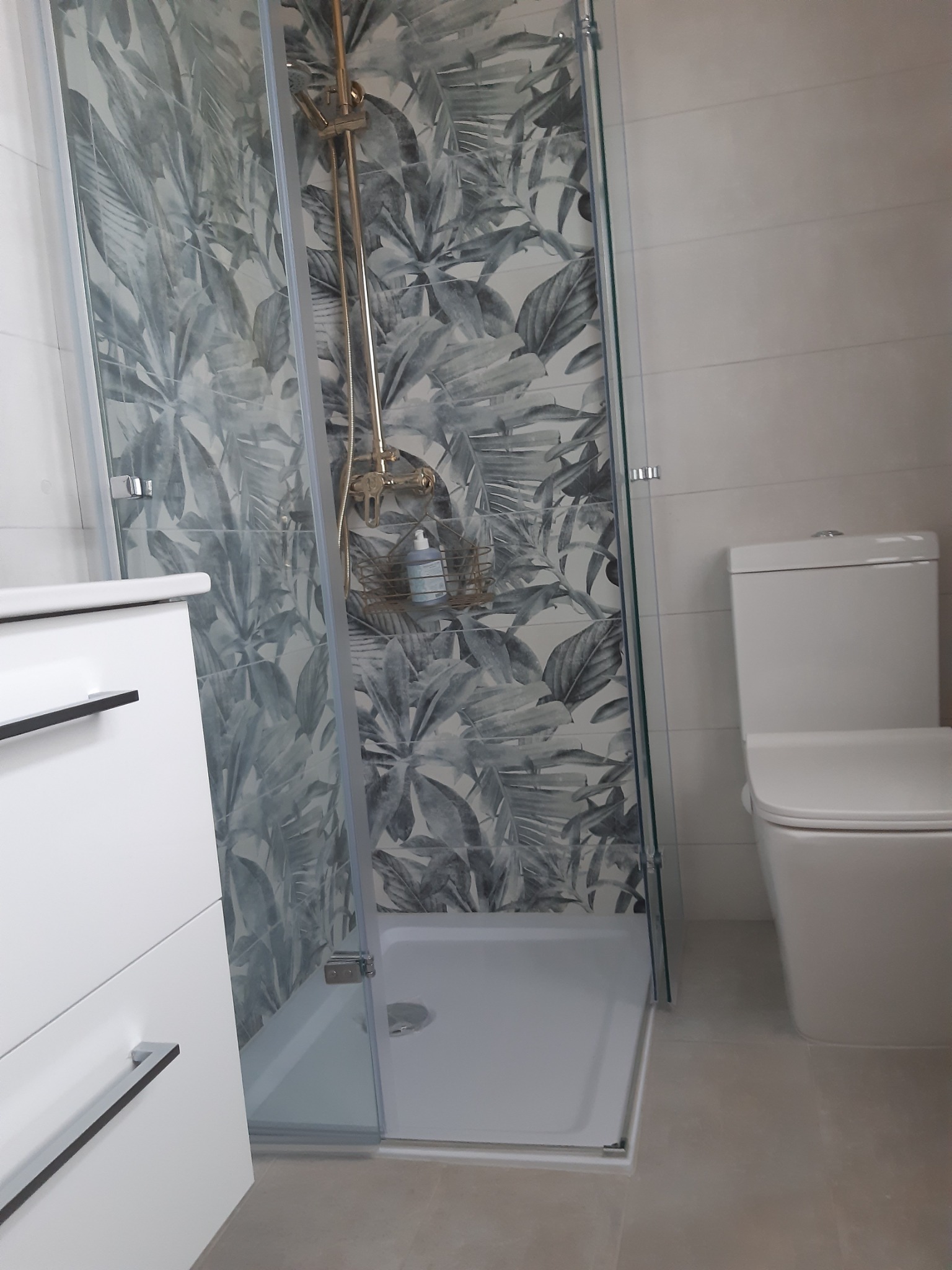 Nowoczesna łazienka po remoncie: kabina prysznicowa z deszczownicą i wzorzystymi płytkami w motywie roślinnym, biała umywalka z szafką i toaleta typu kompakt.