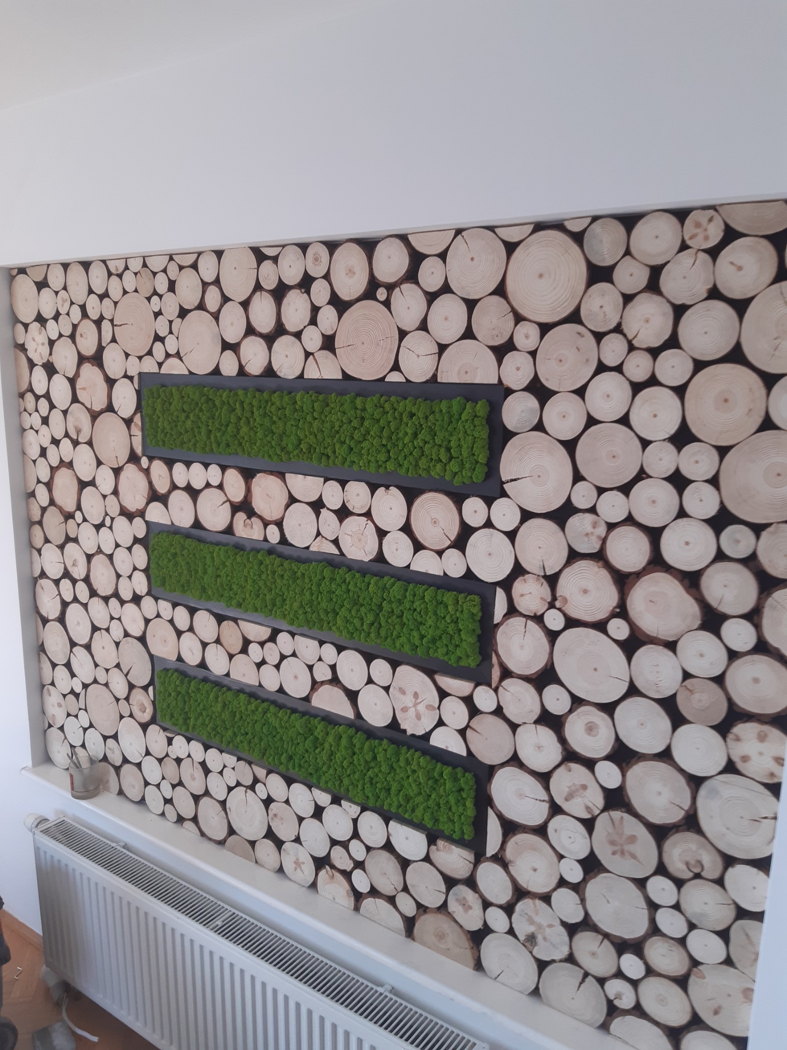 Ściana z dekoracją z drewna i mchu w trzech poziomych pasach, widoczny grzejnik pod spodem.