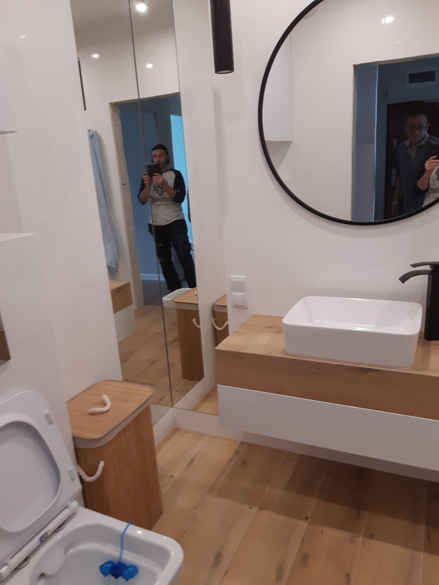 Nowoczesna łazienka z drewnianymi akcentami, białą umywalką nablatową i okrągłym lustrem; widoczne odbicie osoby robiącej zdjęcie w lustrze.