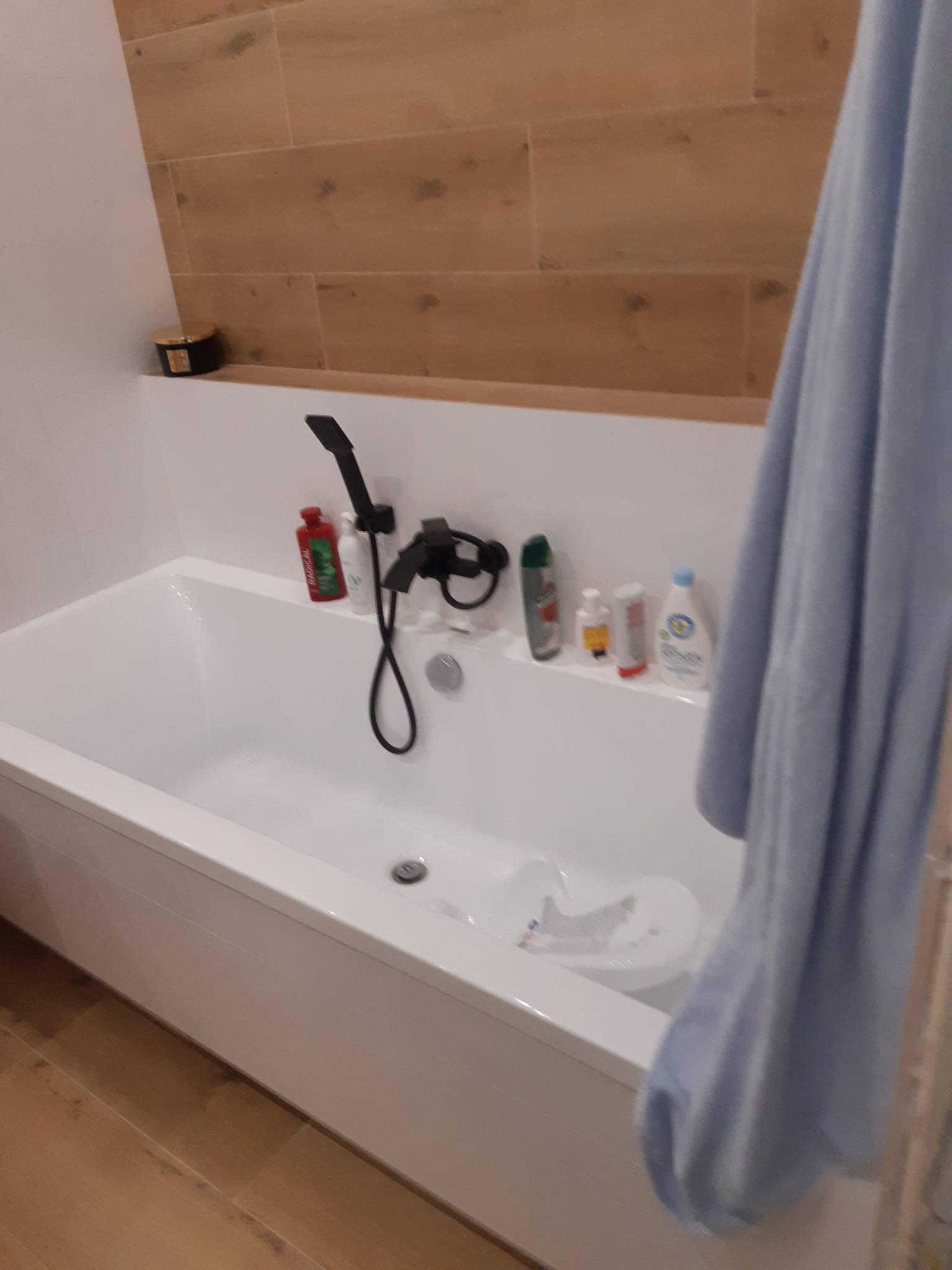 Biała wanna z czarną armaturą, drewnopodobne płytki na ścianie i niebieski ręcznik, widok na półkę z kosmetykami.