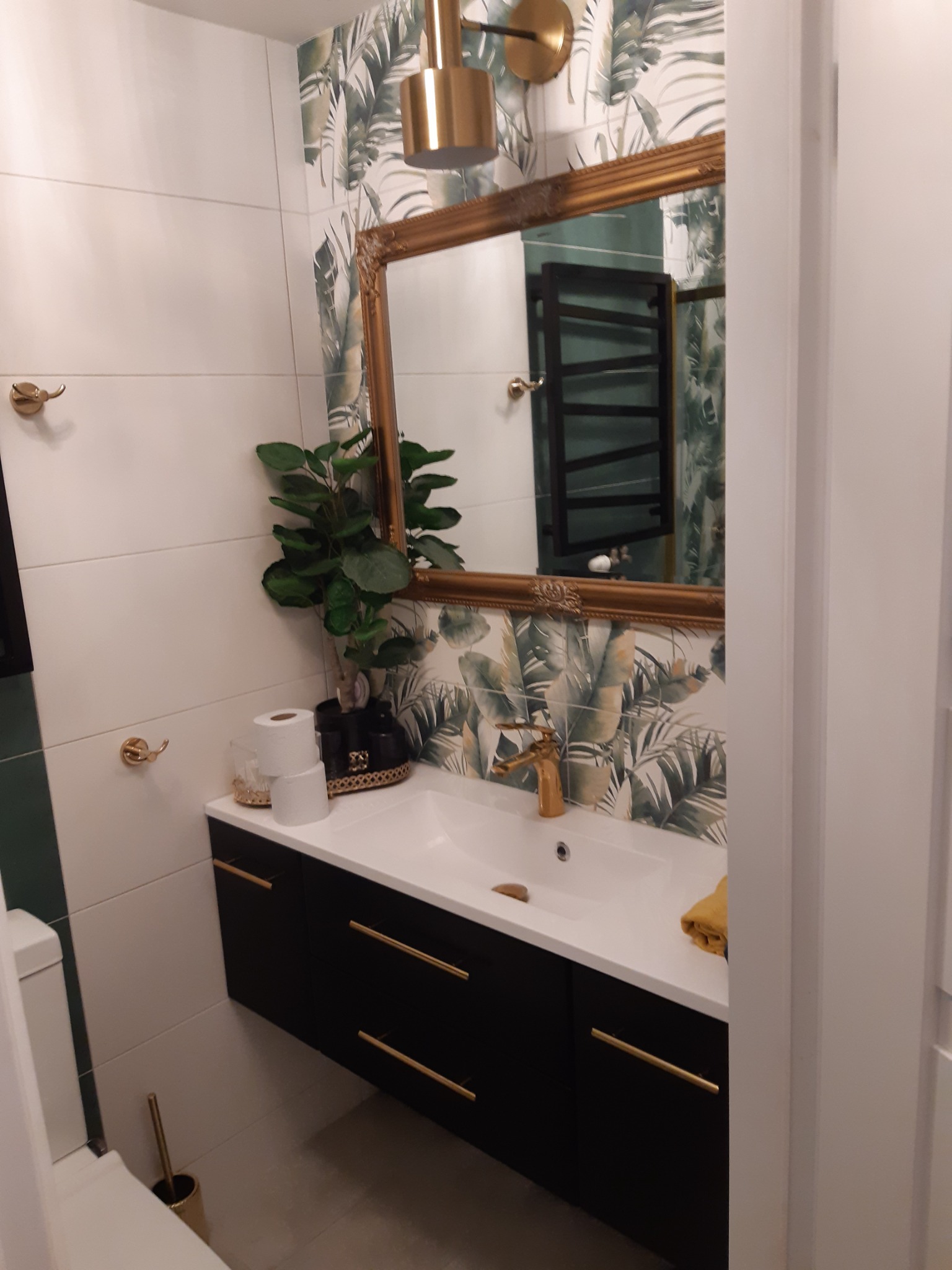 Nowoczesna łazienka z umywalką na szafce w kolorze czarnym, złota armatura, lustro w złotej ramie i tapeta z motywem liści palmowych.