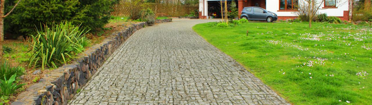 Długa, wybrukowana kostką granitową droga do domu z garażem. Po lewej stronie widoczny murek z kamienia, a po prawej trawnik z kwiatami. W tle zaparkowany szary samochód.