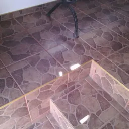 Brązowe płytki podłogowe z wzorem kamienia, wykończenie schod&oacute;w z listwą. Fragment czarnej metalowej nogi mebla w tle.