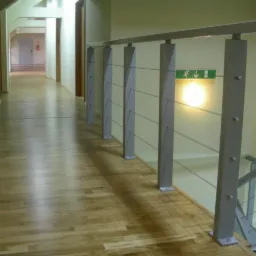Korytarz z drewnianą podłogą i metalową balustradą z linkami, widoczny znak ewakuacyjny.