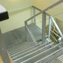 Metalowe schody z kratowymi stopniami i balustradą z metalowych słupków oraz poziomych linek, widok z góry w kierunku schodów.