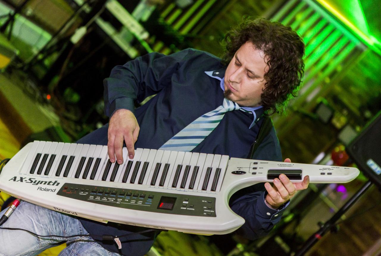 Mężczyzna grający na keytarze Roland AX-Synth, ubrany w niebieską koszulę i niebiesko-biały krawat, oświetlony zielonym światłem scenicznym.