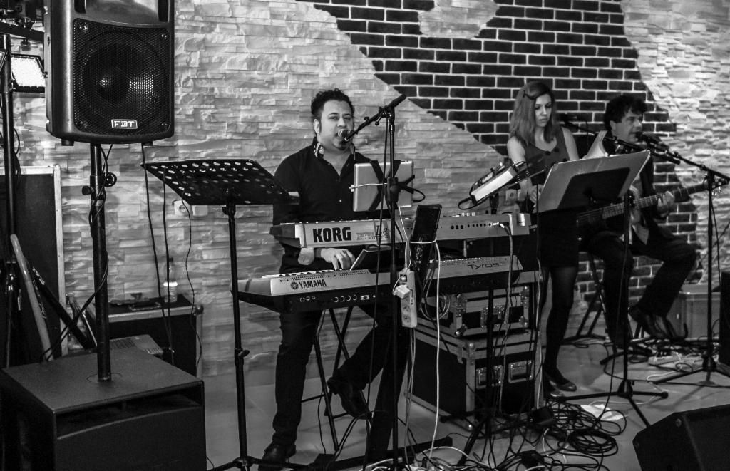 Czarno-białe zdjęcie zespołu podczas występu na żywo, z wokalistą grającym na keyboardzie Yamaha i Korg, gitarzystą i kobietą grającą na keyboardzie Tyros, na tle ściany z cegieł.