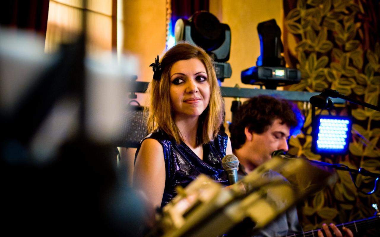 Wokalistka z mikrofonem i gitarzysta podczas występu na scenie z oświetleniem punktowym i dekoracyjną ścianą w tle.